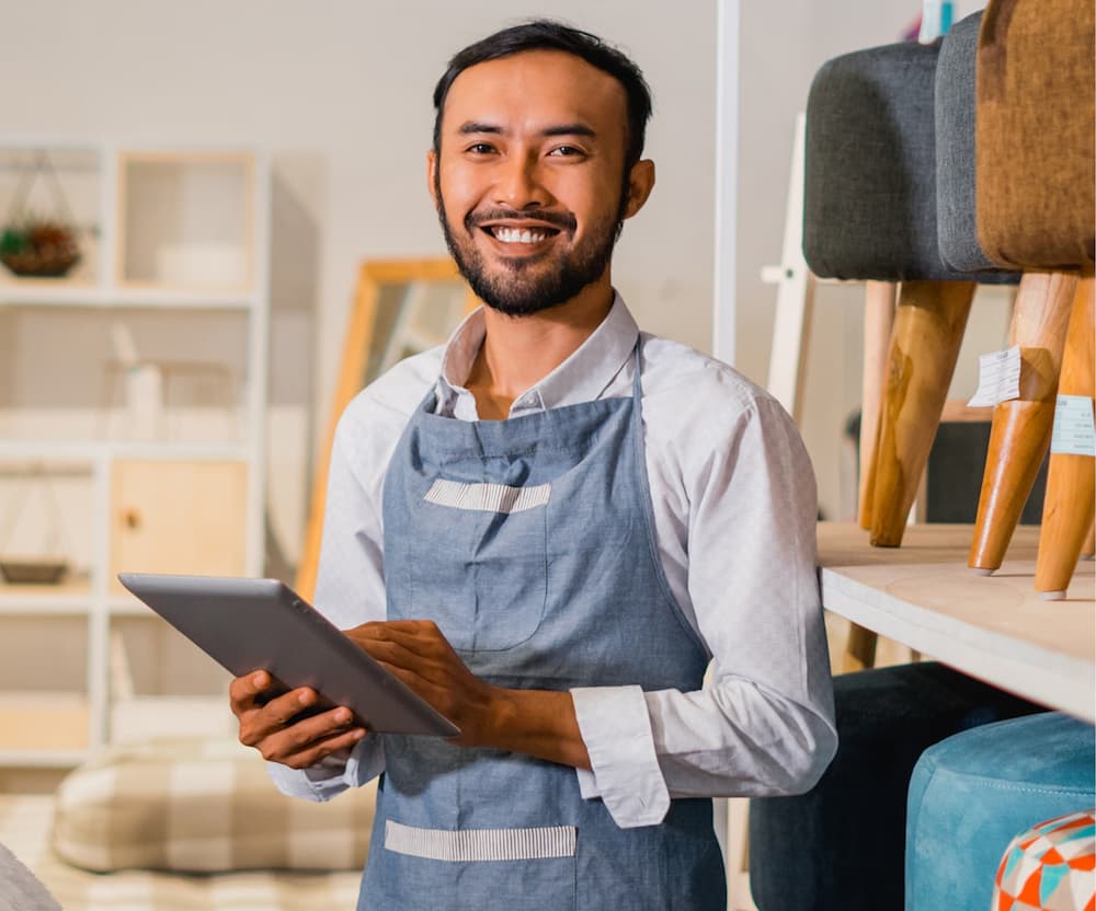 Hombre con delantal sostiene una tableta, sonriendo mientras mira la pantalla, en un ambiente de cocina.